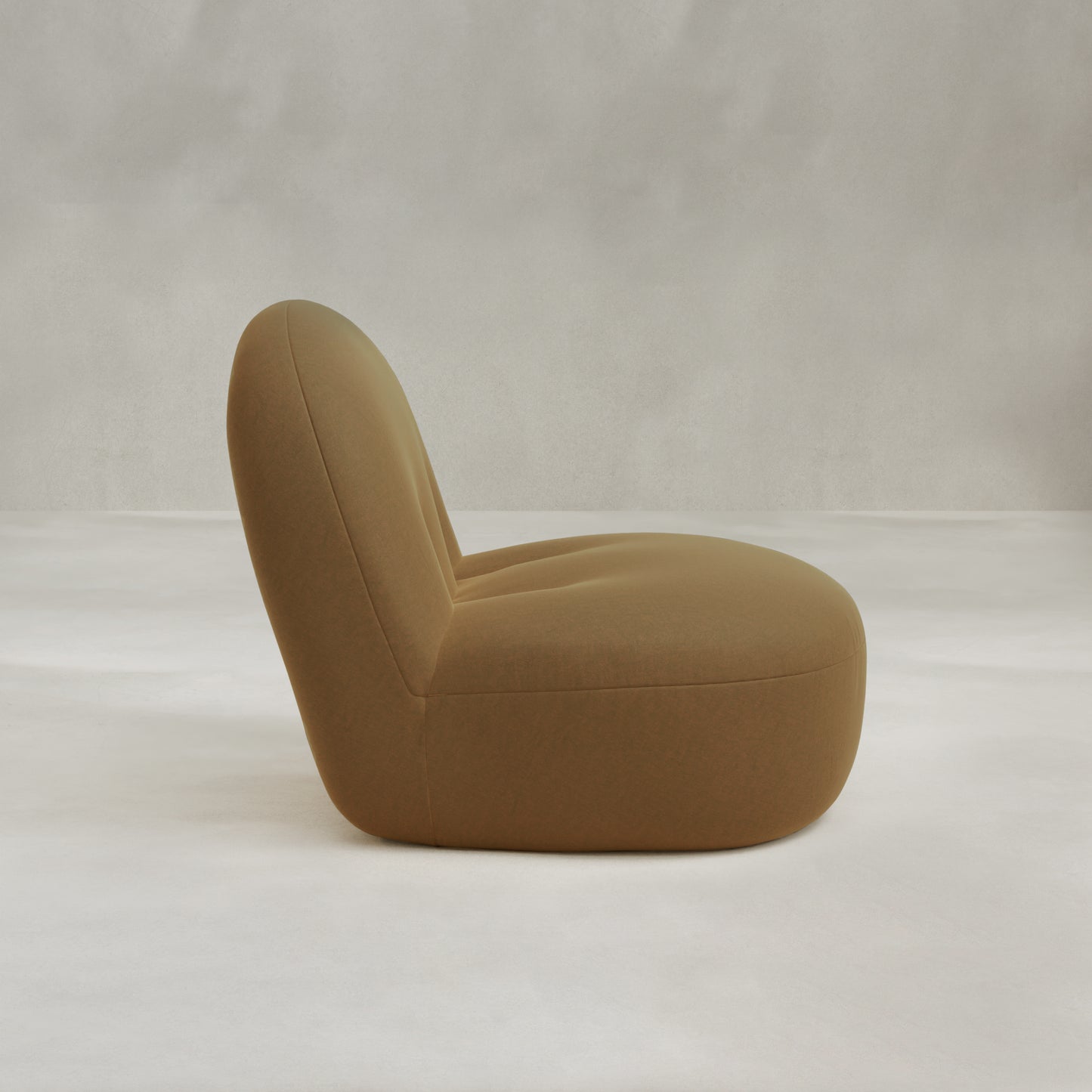 The Colette Chair, a modern armless lounge with a rounded back and seat, is upholstered in light brown fabric and features plush cushions. It sits on a light neutral floor against a plain gray wall.