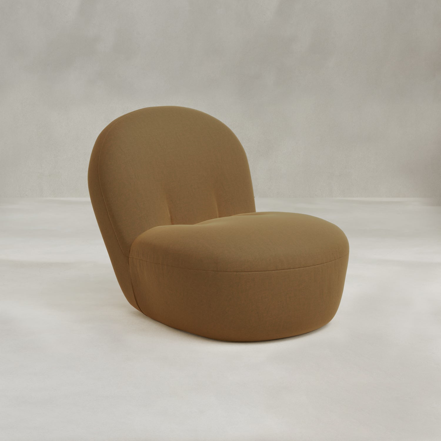 The Colette Chair is a modern, armless lounge chair with a curved backrest and seat, upholstered in richly textured light brown fabric, placed on a neutral surface against a softly textured background.