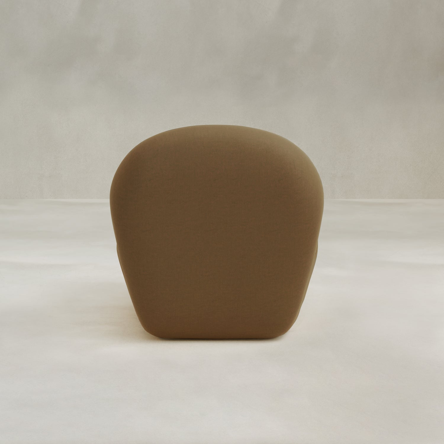 The Colette Chair, a minimalist design with brown rounded form and smooth upholstery, is shown from the back on a light floor against a softly textured pale background.