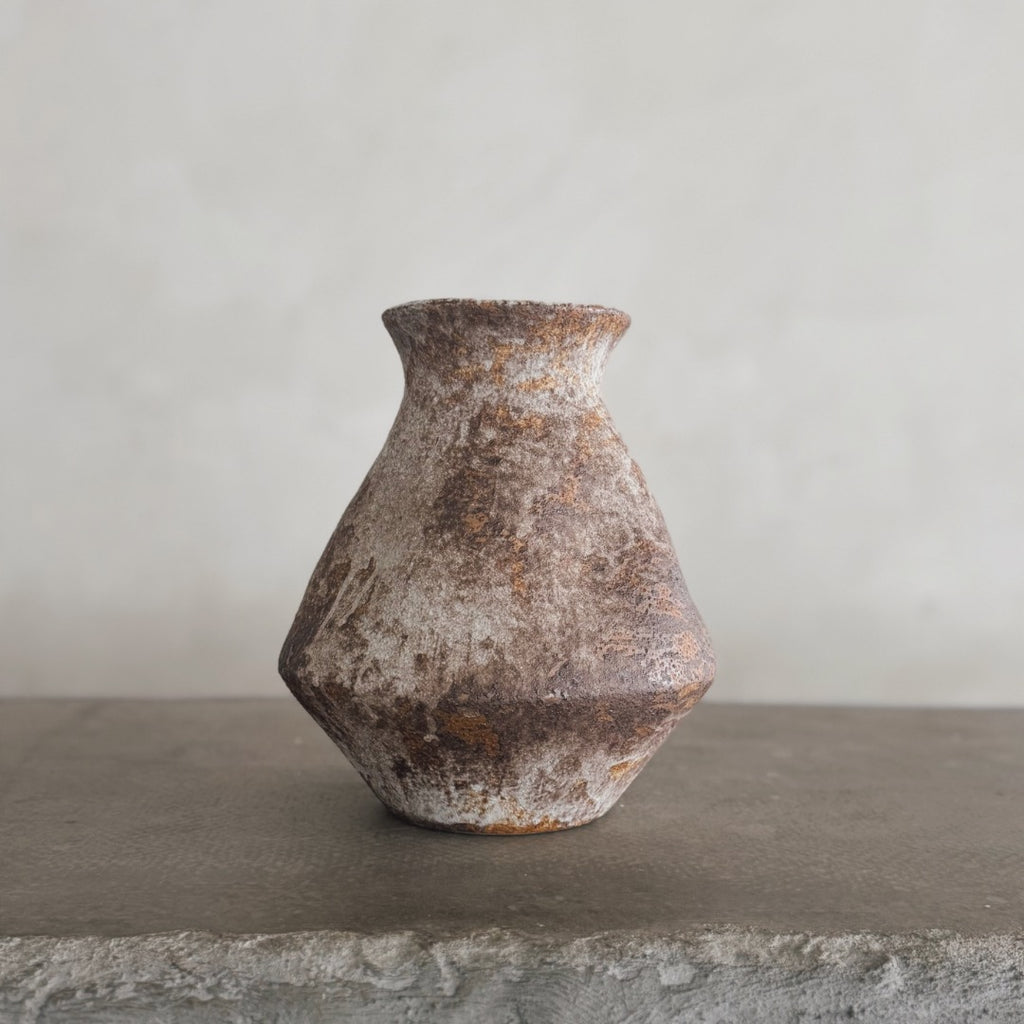The Nami vase, featuring a rustic, weathered ceramic look and textured surface, sits on a rough gray stone ledge against a light gray backdrop—ideal for bringing farmhouse decor charm to your space.