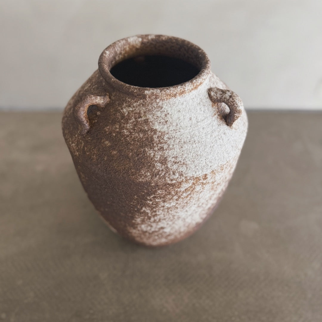 The Yuki vase is a rustic, brown and white ceramic piece with two small handles near the top, displayed on a neutral surface against a blurred background.
