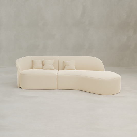 The Marlow Curved Modular Three Seater Sofa, upholstered in light beige velvet with a modern curved design, includes three matching square pillows and is set against a neutral gray wall on a light gray floor.