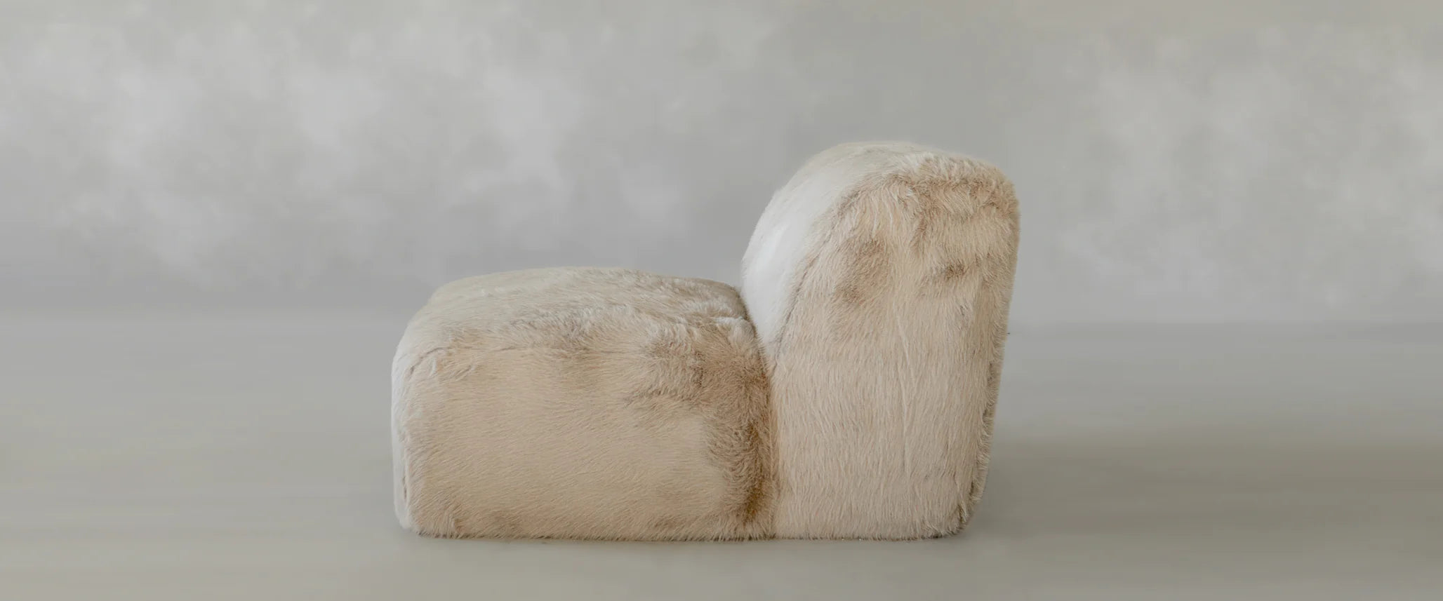 The Yeti One Seater Modular, a modern armless sofa with a low back, is fully upholstered in beige faux fur and set against a matching gray wall—ideal for any contemporary living space.