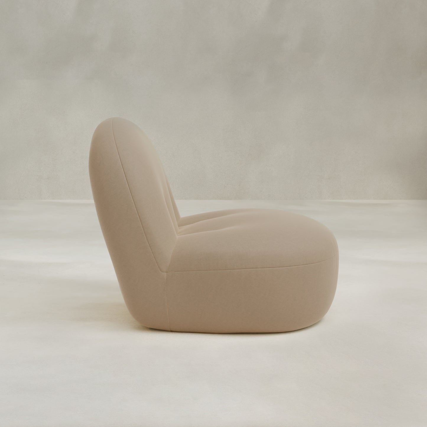 The Colette Chair, a modern armless lounge seat featuring plush beige cushions and a rounded minimalist design, is displayed in profile against a light neutral background.