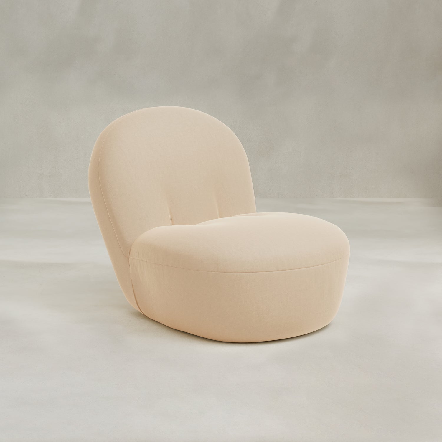 The Colette Chair is a modern, armless lounge chair with light beige upholstery, a rounded backrest, and plush cushions, all set against a neutral, textured background for contemporary style.