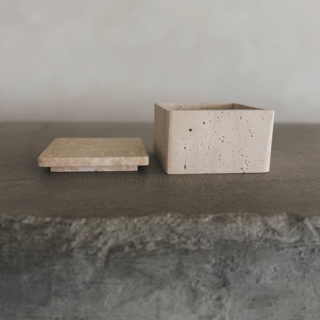 The Celeste Vault with Lid, a square beige stone box featuring a matching flat lid, sits open on a textured gray stone surface. The minimalist design and natural materials create a clean, elegant look.
