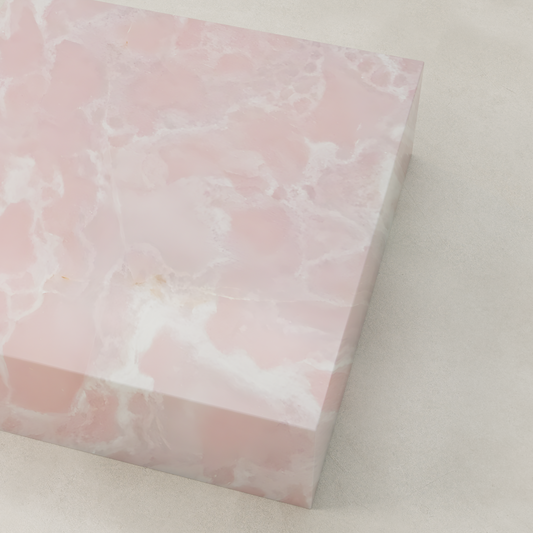 The Cora Onyx Coffee Table XL is a large rectangular piece featuring glossy pink onyx with white veining, a soft cloudy texture, and a polished finish. It sits elegantly on a light-colored, smooth surface.