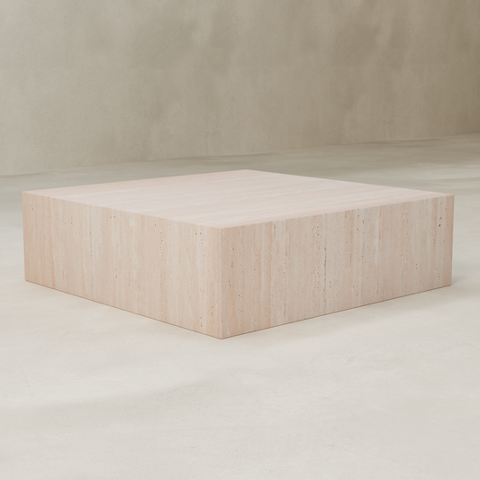 The Alessia Coffee Table XL, a large square travertine plinth with a minimalist block design and natural stone texture, sits on a smooth neutral floor against a plain light wall, creating a striking modern statement.