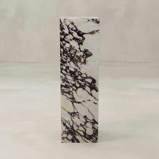 The Calacatta Tall Tower Plinth features a tall, rectangular profile with dramatic dark brown and black veining, standing upright on a light, textured surface against a neutral background.