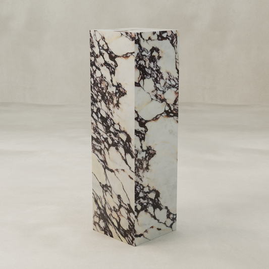 The Calacatta Tall Tower Plinth features a tall, rectangular form with a white base and striking dark brown marble veining, standing on a light, textured floor against a neutral background to highlight its luxurious hues.