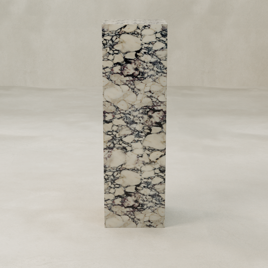 The Azurine Tall Tower Plinth features marble elegance with a cream background and dark, intricate veins. It stands on a smooth, light gray surface against a plain, textured wall.