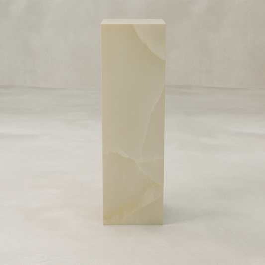 The Lunelle Onyx Tall Tower Plinth features smooth, cream-toned stone with a milky hue and subtle golden veining, standing tall and rectangular on a light, textured surface against a neutral backdrop.