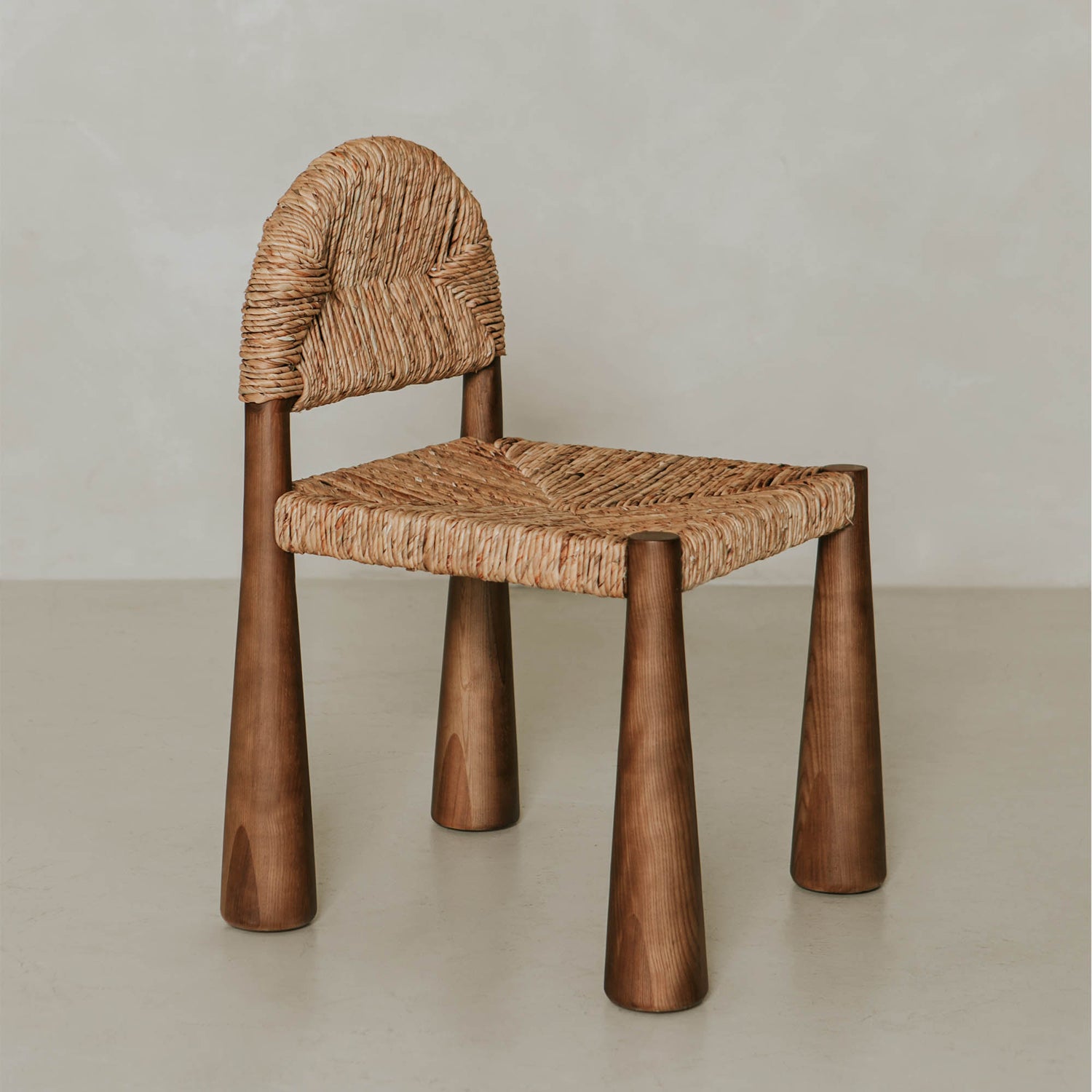 The Renè + Bardo Dining Set features a modern chair with a woven, straw-like seat, rounded backrest, and four wide, tapered wooden legs, shown against a plain light background.