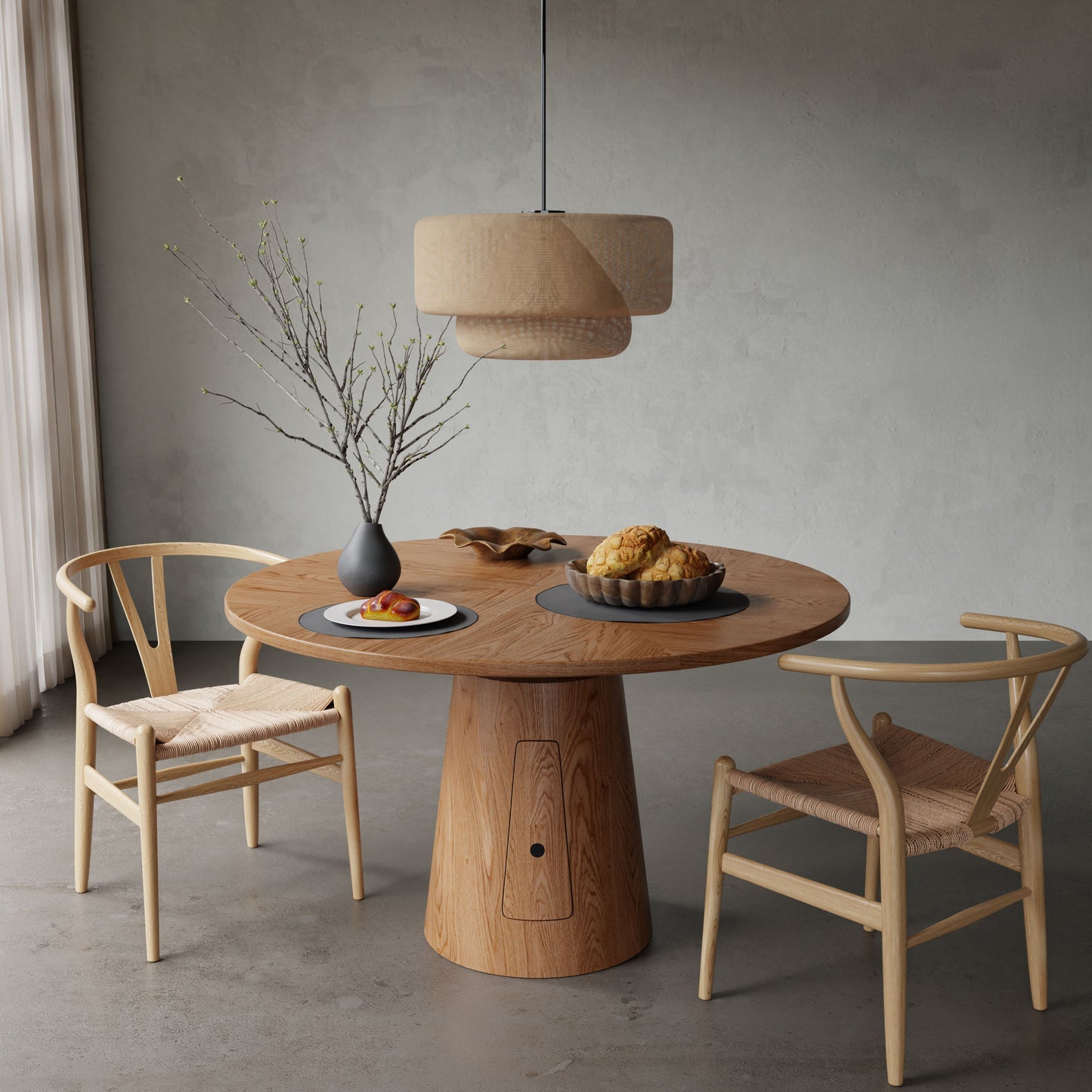 The Nola + Elsa Dining Set, featuring a round wood table and two woven seat chairs, is styled with a vase of branches, a pastry plate, and a fruit bowl under a pendant lamp in a softly lit, neutral-toned room.