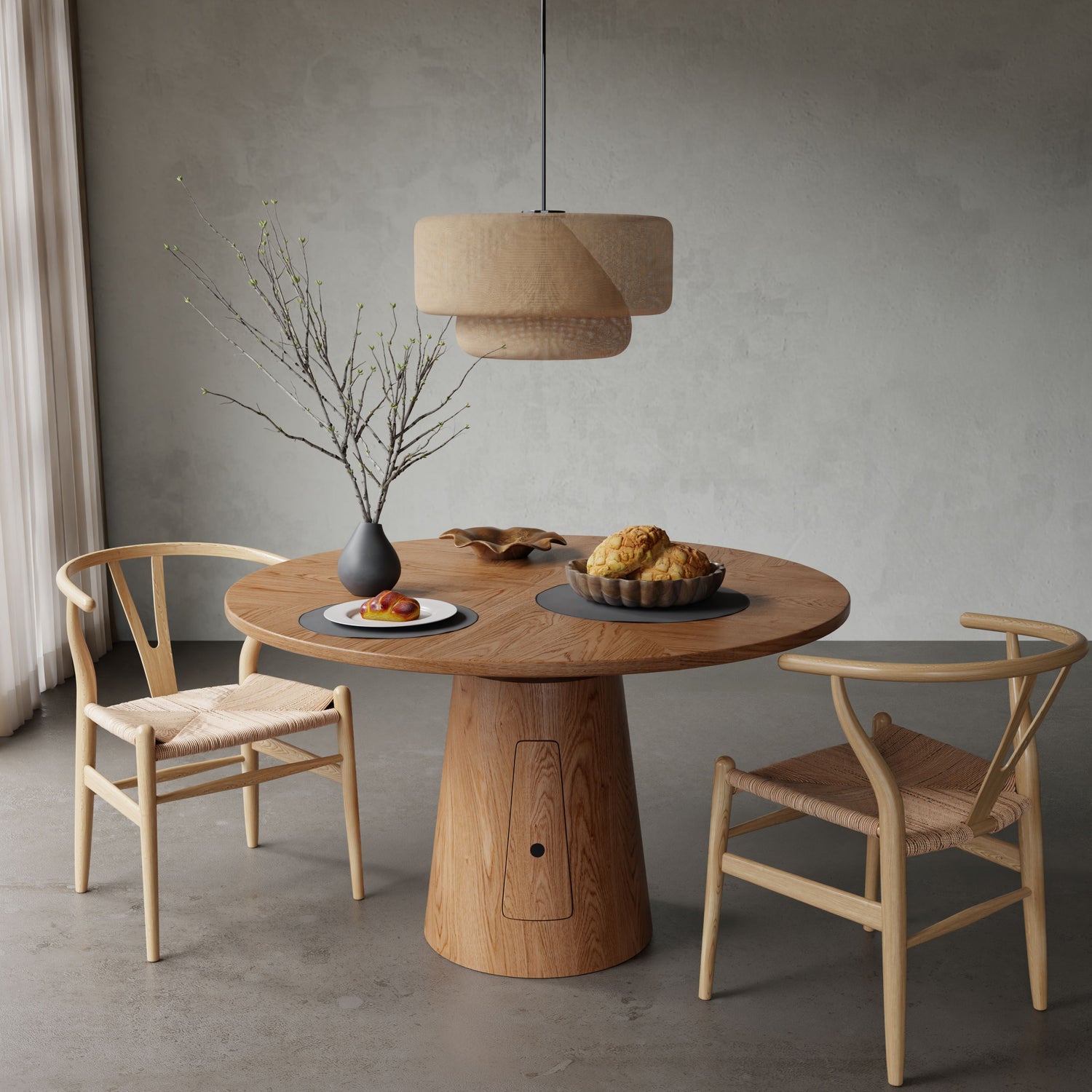 The Nola + Elsa Dining Set, featuring a round wood table and two woven seat chairs, is styled with a vase of branches, a pastry plate, and a fruit bowl under a pendant lamp in a softly lit, neutral-toned room.