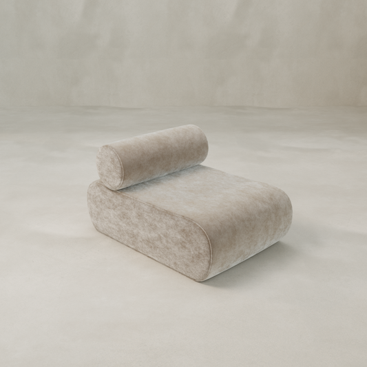 The Nuage Modular Lounge Chair features a modern, minimalist design with a sculptural silhouette and cylindrical bolster backrest, upholstered in soft, velvety fabric, set against a simple light-colored background.