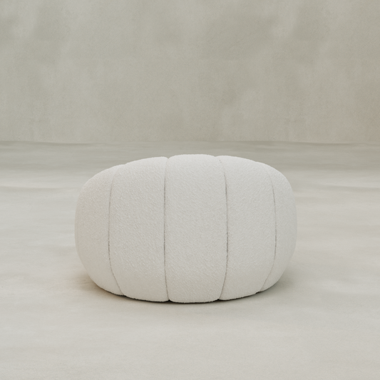 A round, white, textured pouf with vertical seams sits on a smooth, light-colored floor against a neutral backdrop, perfect for pairing with the Penelope Chair to create a cozy reading nook.