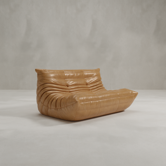 The Rolo Lounge Loveseat, in modern tan leather with a low profile and deep, ruched cushions, brings mid-century flair to light floors and a plain gray wall.