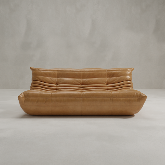 The Rolo Lounge Three Seater, a modern tan leather sofa with tufted cushions and a low profile, features subtle mid-century design touches and stands out against a light-colored wall in a minimalist space.