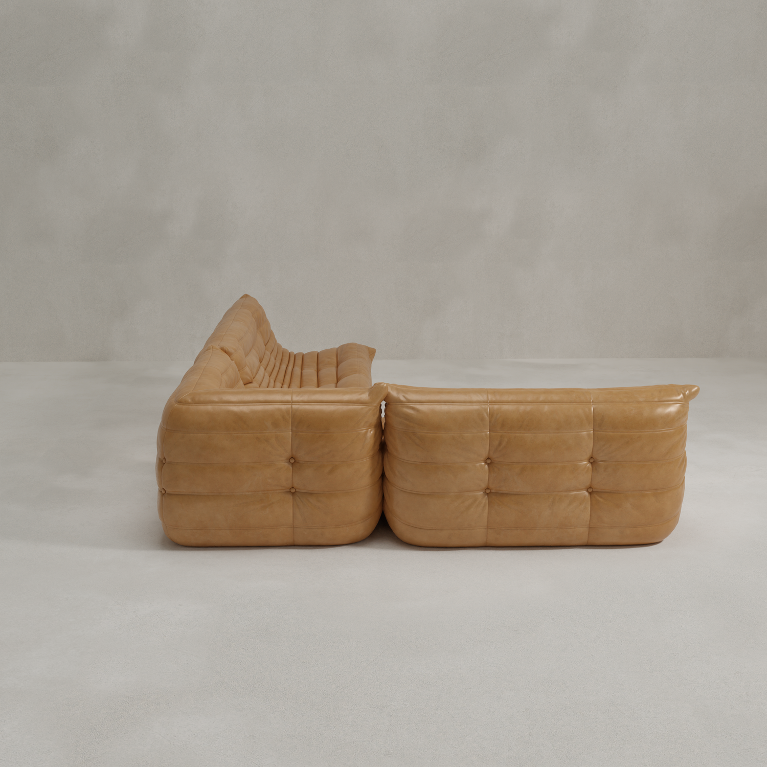 The Rolo Lounge Three Piece Sofa, with plush tan leather tufted cushions and rounded modular design, sits on a light gray floor against a textured gray wall, evoking the comfort and style of a 70s-inspired lounge chair.