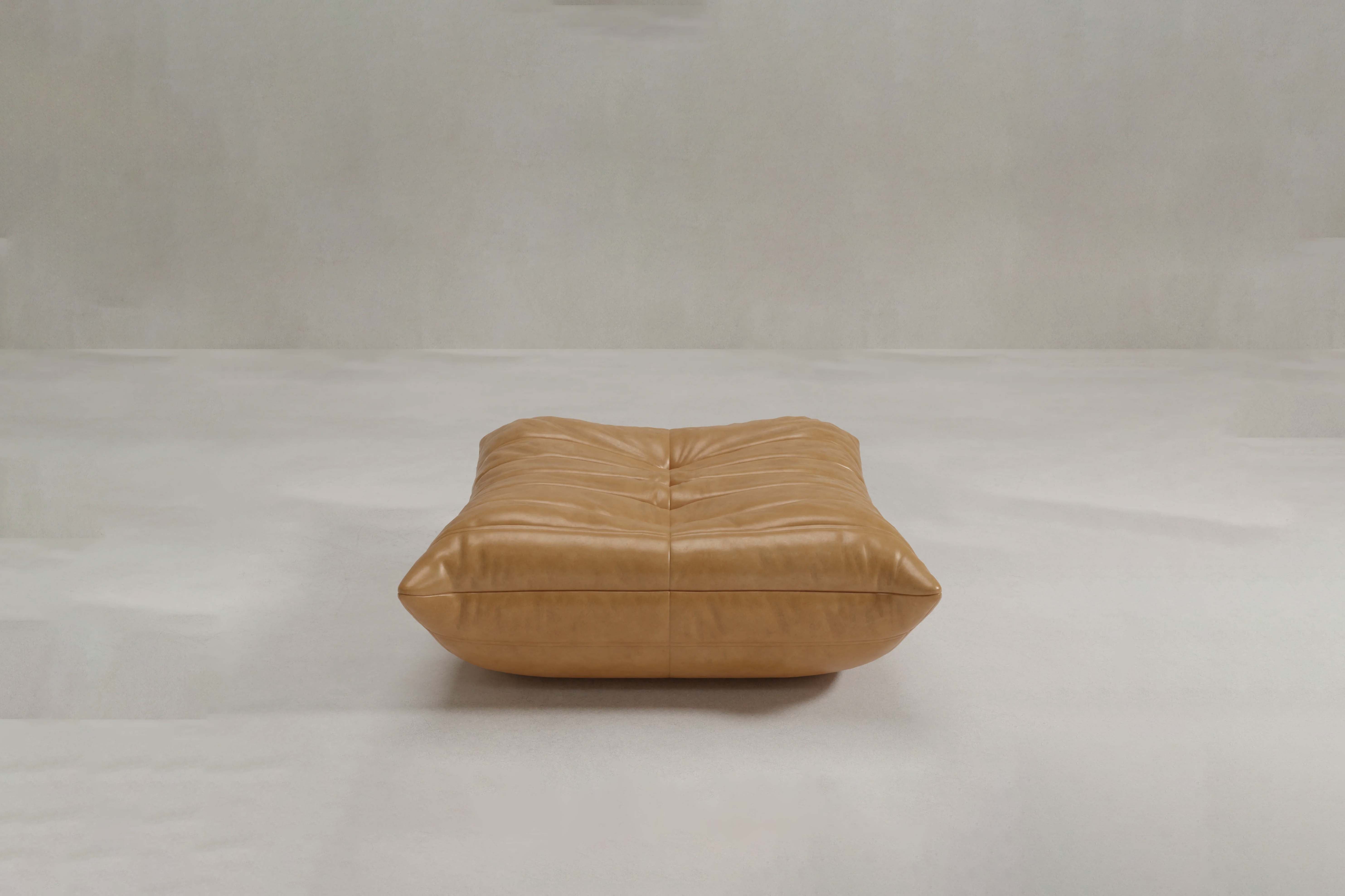The Rolo Ottoman is a large, square tan leather floor cushion with soft horizontal tufted seams, perfect as a versatile ottoman or extra seating, shown here on a light gray surface.