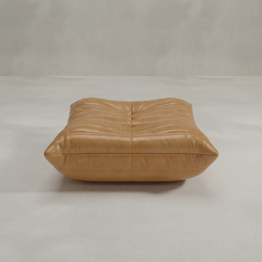 The Rolo Ottoman, a spacious tan leather footstool with tufted and ruched details, offers extra seating or functions as a versatile ottoman in an empty light gray room.