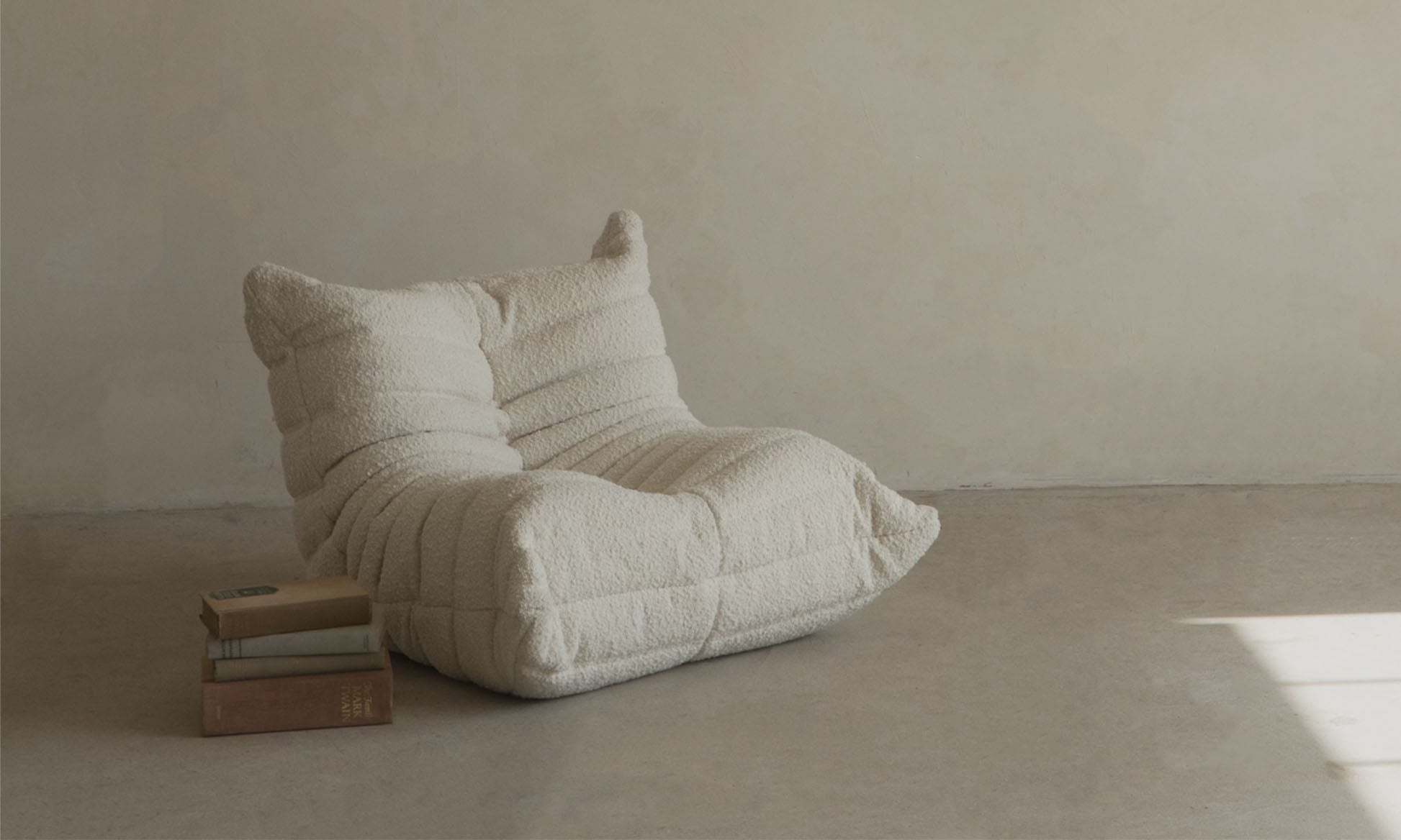 The cream Mini Rolo Lounge Chair, with its cozy textured design, sits on a concrete floor beside books and a beige wall—an ideal choice for a minimalist playroom or child’s space.