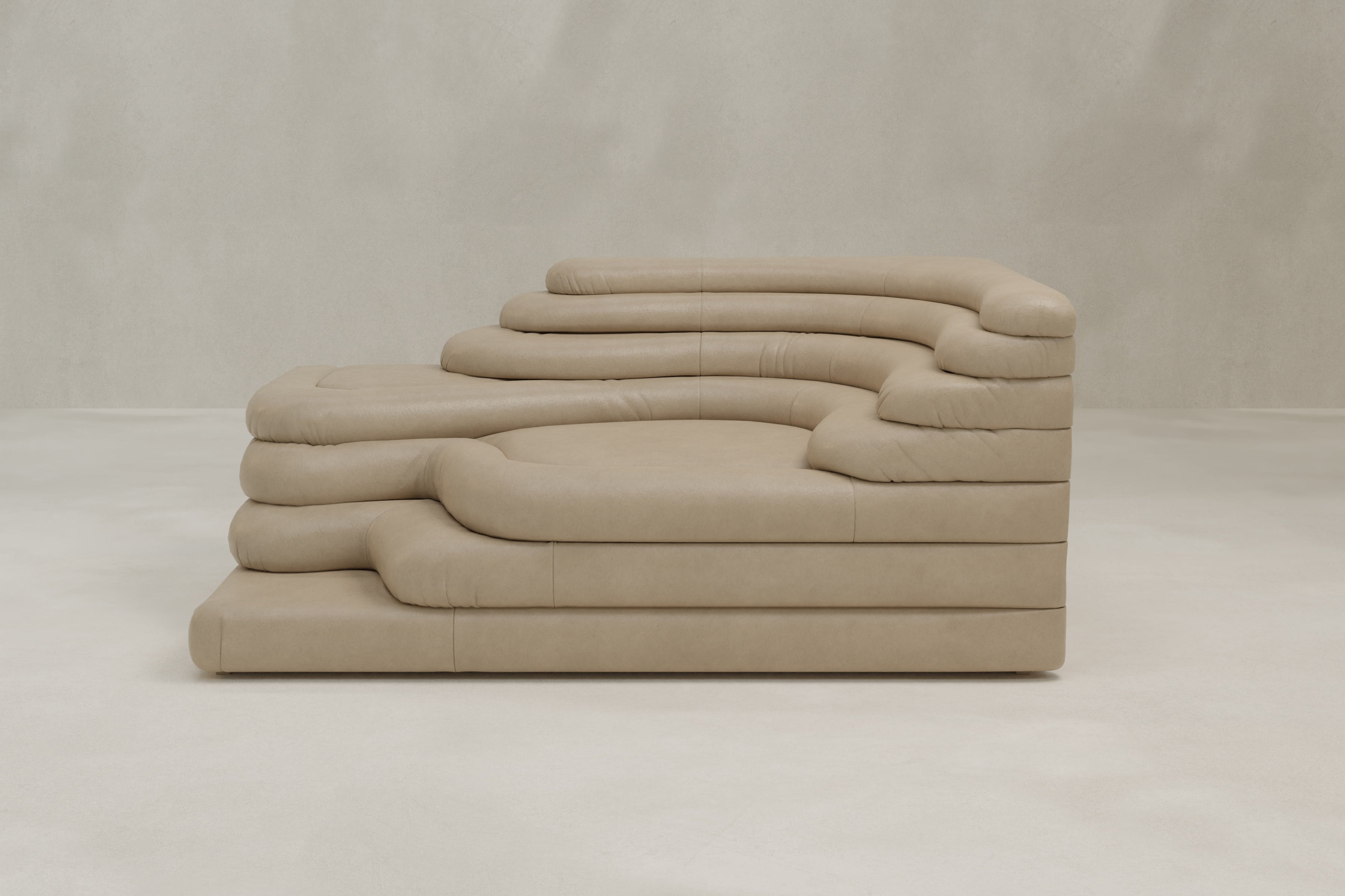 The Cascata Sofa Left Arm is a sculptural beige leather modern sofa featuring layered, wave-like cushions and a sleek design, perfectly complementing a neutral room with plain light walls and flooring.