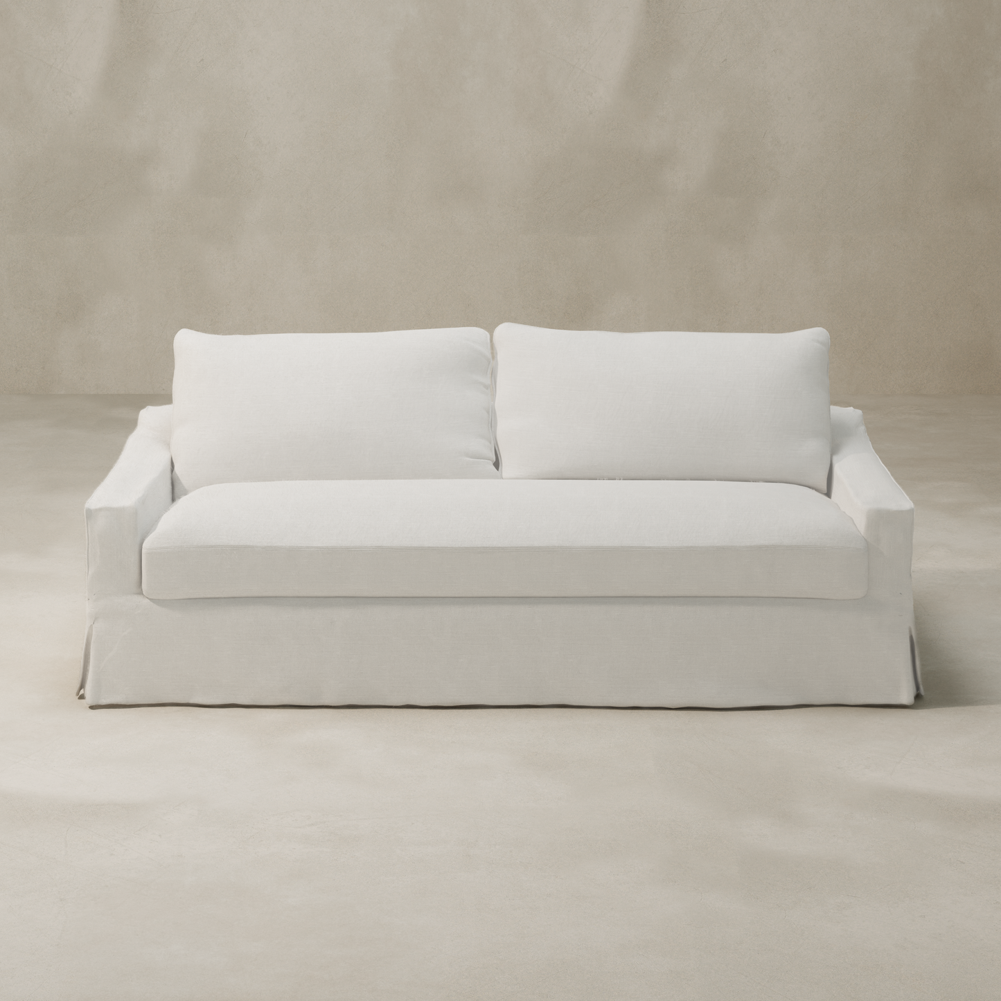 A modern Woolsey Sofa with a skirted base and natural-toned fabric is placed in a minimalist room featuring light beige walls and flooring.
