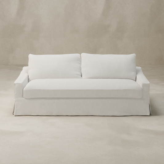 A modern Woolsey Sofa with a skirted base and natural-toned fabric is placed in a minimalist room featuring light beige walls and flooring.