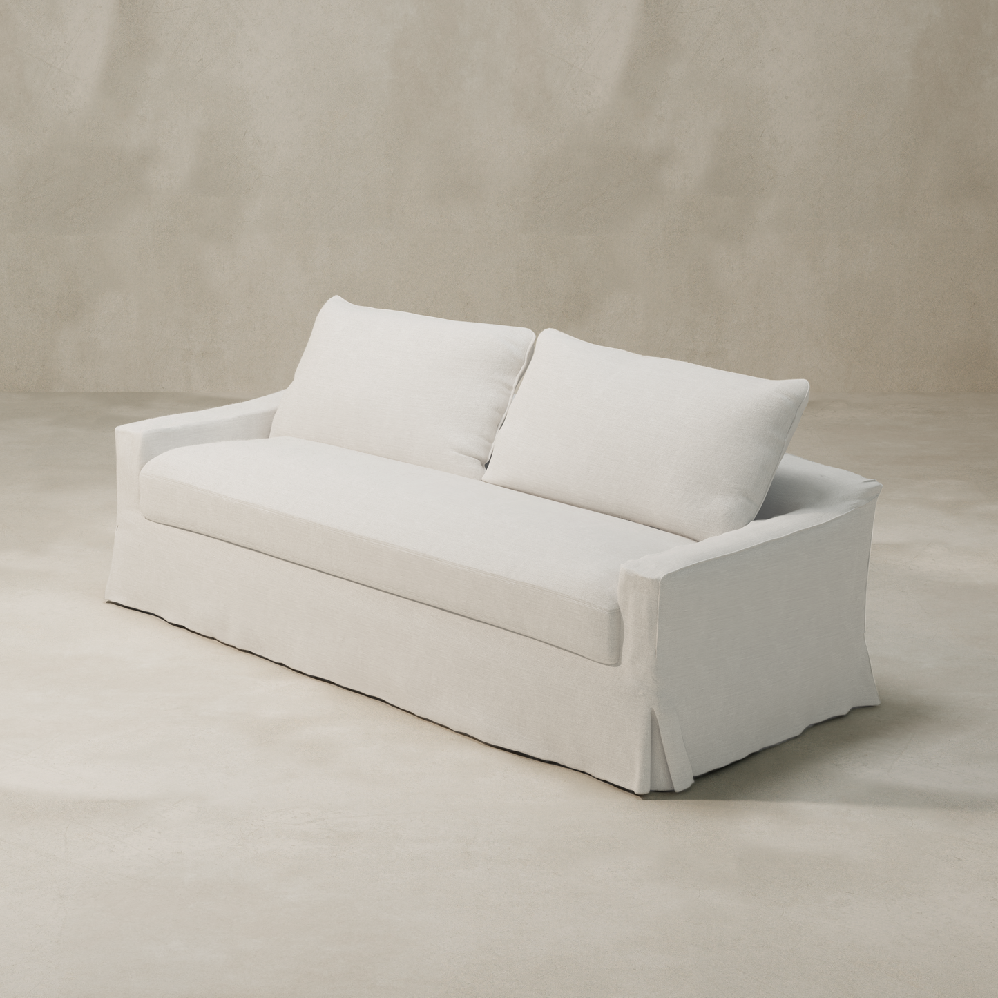 The Woolsey Sofa is a modern white piece featuring two large back cushions and a tailored, skirted base in natural-toned fabric, set against a plain beige background.