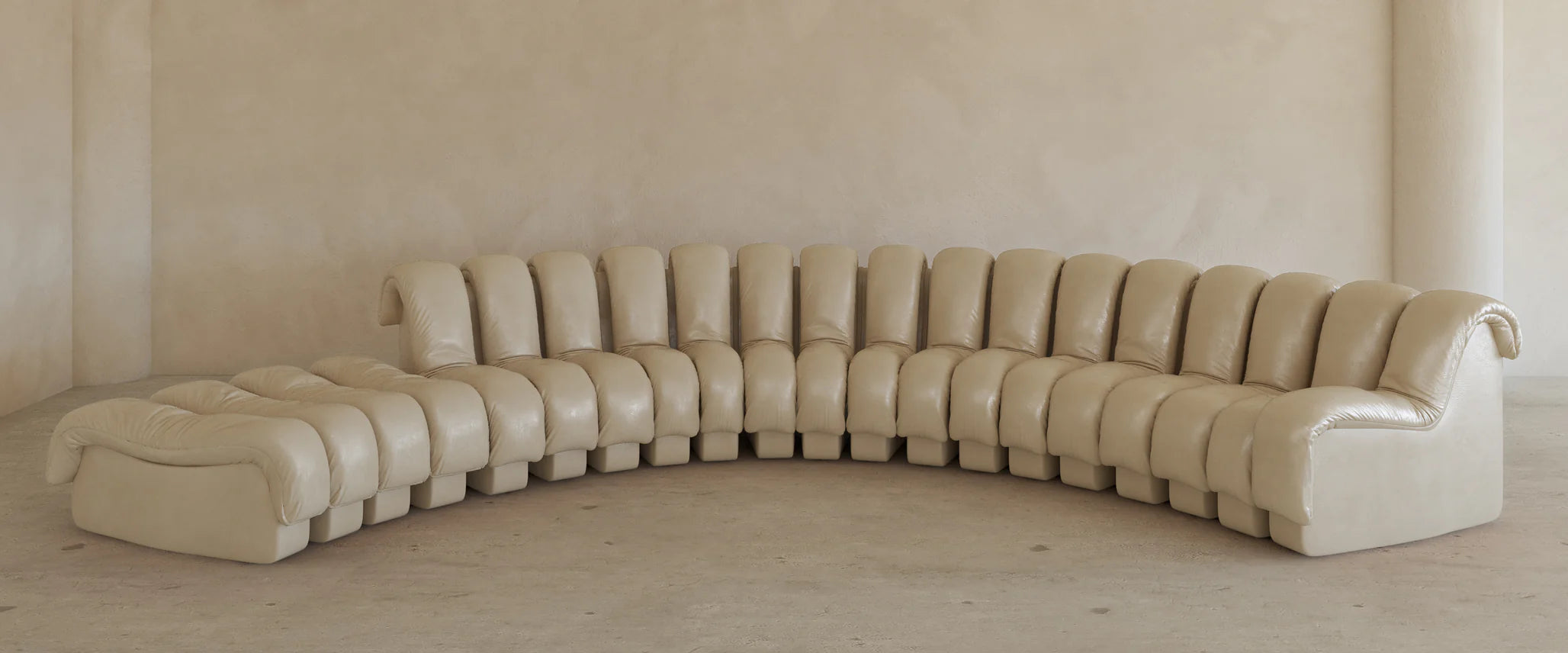 The Serpenti Sofa 15 Piece With Ottoman Rental is a large, curved beige leather modular system with tufted cushions and addable armchair elements for flexible seating, set against a plain light wall in a minimalist room.