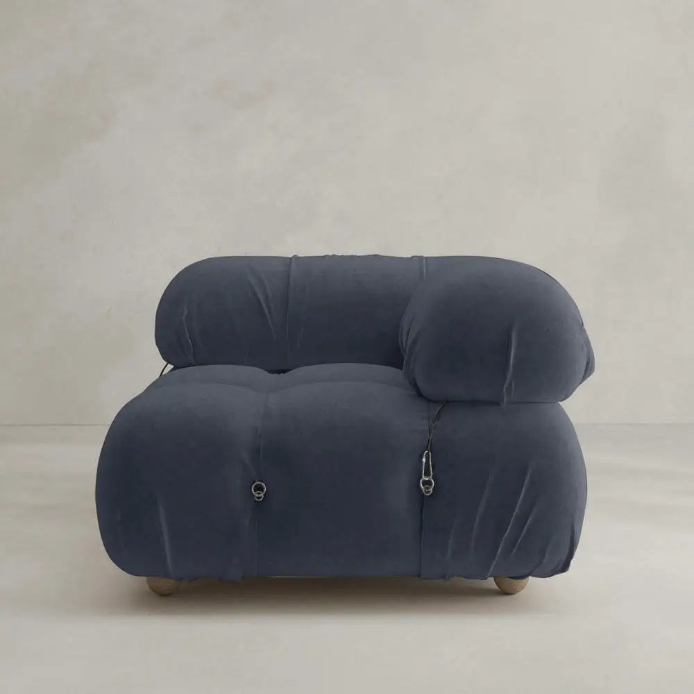 The Belmont Sofa Left Armrest features dark blue upholstery, rounded cushions, and visible metal fasteners, highlighting classic style in a minimalist room with light-colored walls and flooring.