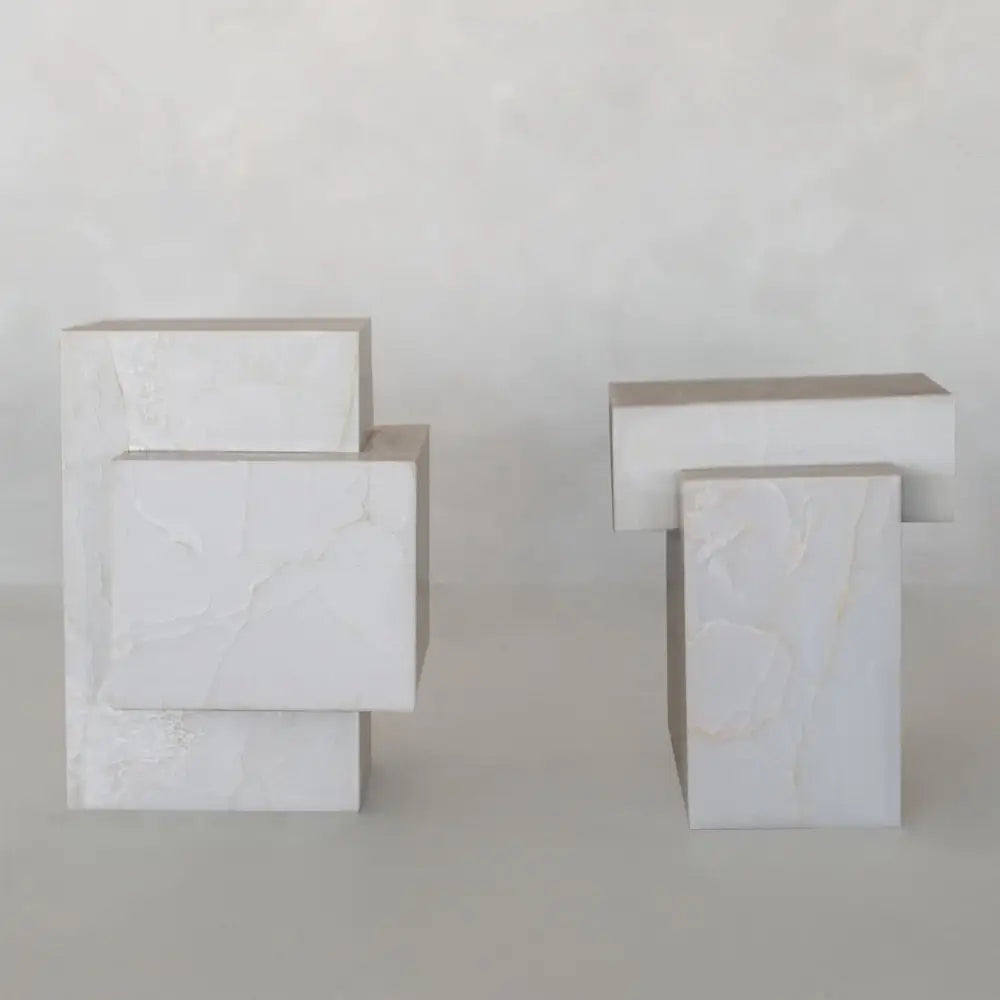 The Bianca Plinth Set features two geometric sculptures of intersecting rectangular and square blocks, crafted from white onyx plinths with subtle veining for a modern, elegant look against a neutral backdrop.