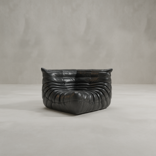 The Rolo Lounge Corner Sofa, a modern black leather piece with deep ruched cushioning and 70s-inspired design, sits on a smooth light gray floor against a plain, textured gray wall.