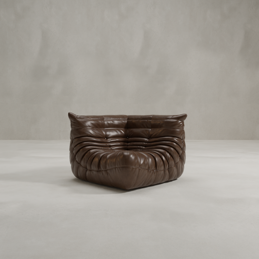 The Rolo Lounge Corner Sofa, with its modern low-profile design and deep brown tufted leather upholstery inspired by mid-century style, sits on a minimalist concrete floor against a plain gray wall.