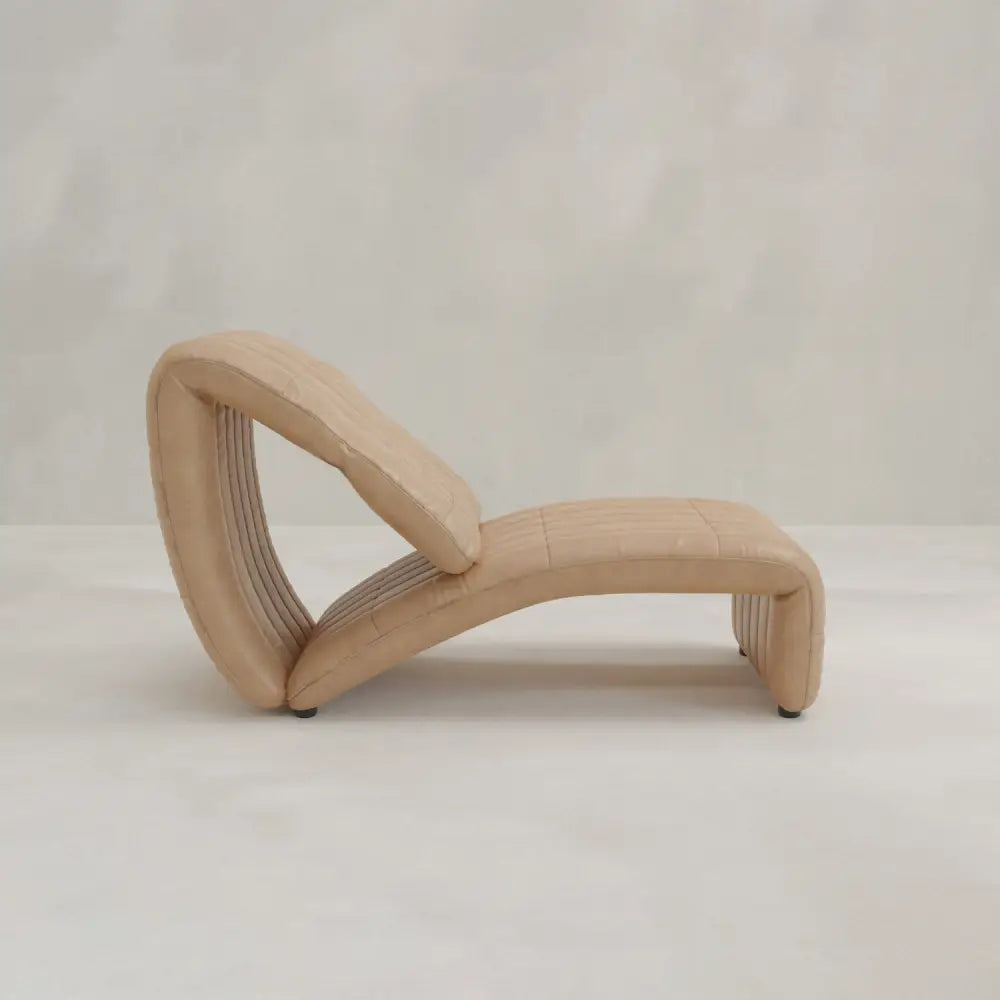 The Hillier Chair features a sculptural, curved design in premium vintage leather, offering smooth lines and a minimalist look. Its subtle reclining adds comfort, complemented by a neutral-toned floor and matching background.