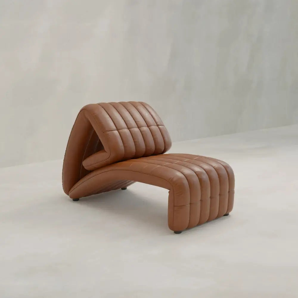 The Hillier Chair features sculptural curves and ribbed premium vintage leather upholstery, shown here on a light floor with a plain, textured beige wall backdrop.