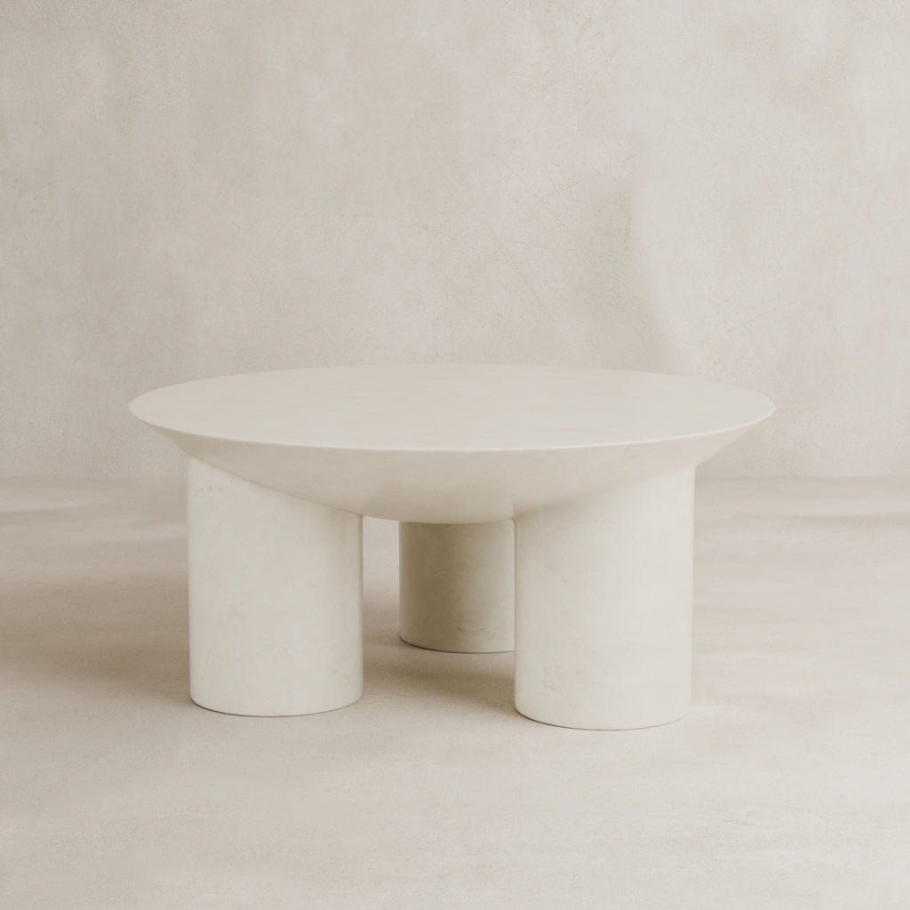 The Luna Coffee Table showcases modern minimalist elegance with thick tubular legs and a smooth off-white fiberglass finish, complemented by a neutral, textured backdrop.