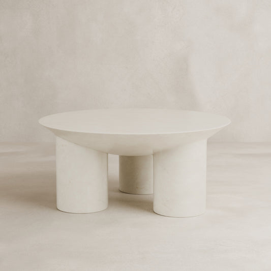 The Luna Coffee Table showcases modern minimalist elegance with thick tubular legs and a smooth off-white fiberglass finish, complemented by a neutral, textured backdrop.