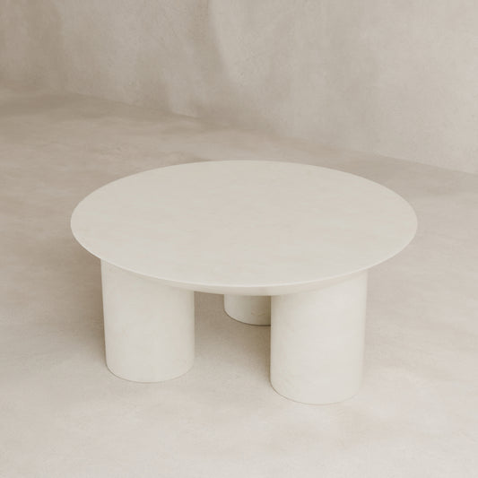 The Luna Coffee Table, featuring a minimalist round fiberglass top and three tubular legs, is showcased against a light beige textured background, radiating modern elegance.