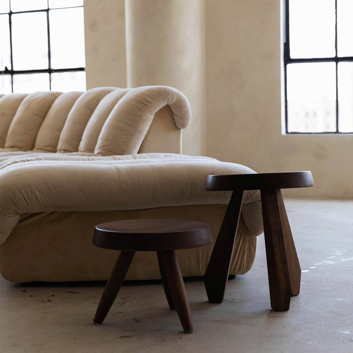 A modern living room showcases a cream plush sofa with curved backrest and two Amara and Athena Stools as tables, all set on a light floor near large industrial-style windows.