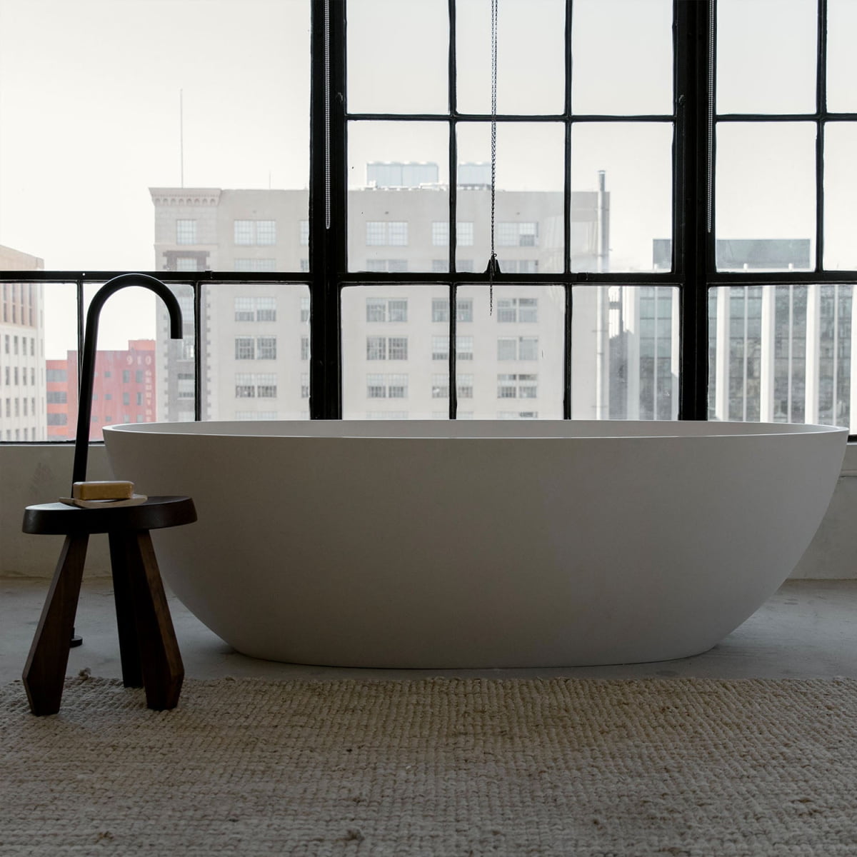 A modern freestanding white bathtub sits on a textured rug beside the Amara Tall Stool holding soap, in front of large windows overlooking city buildings.