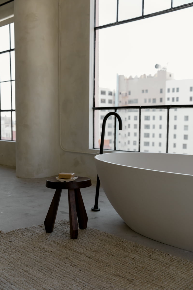 A modern bathroom features a freestanding white bathtub, a black faucet, and the Amara Tall Stool with a tripod base holding soap, set beside large windows overlooking city buildings.