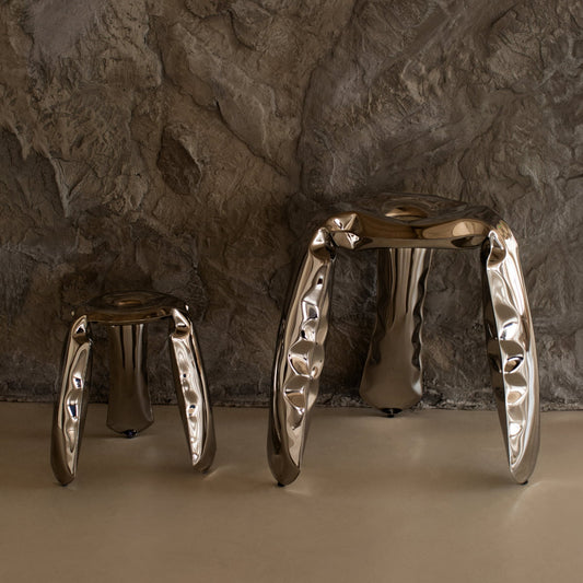 Two metallic Araknid stools, each with three legs and a shiny crumpled look, stand on a beige floor by a textured stone wall. Their reflective surfaces make the Araknid Stool Set a striking seating choice.