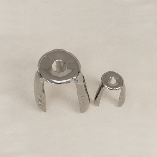 The Araknid Stool Set features two abstract, metallic stools with three legs and organic shapes. Each stool has a round top with a central hole, offering unique seating experiences on a light, textured surface.