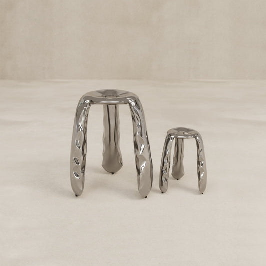The Araknid Stool Small features an organic, wavy design with a shiny metallic finish and three ergonomic legs, offering comfort and sophistication against a plain beige background.