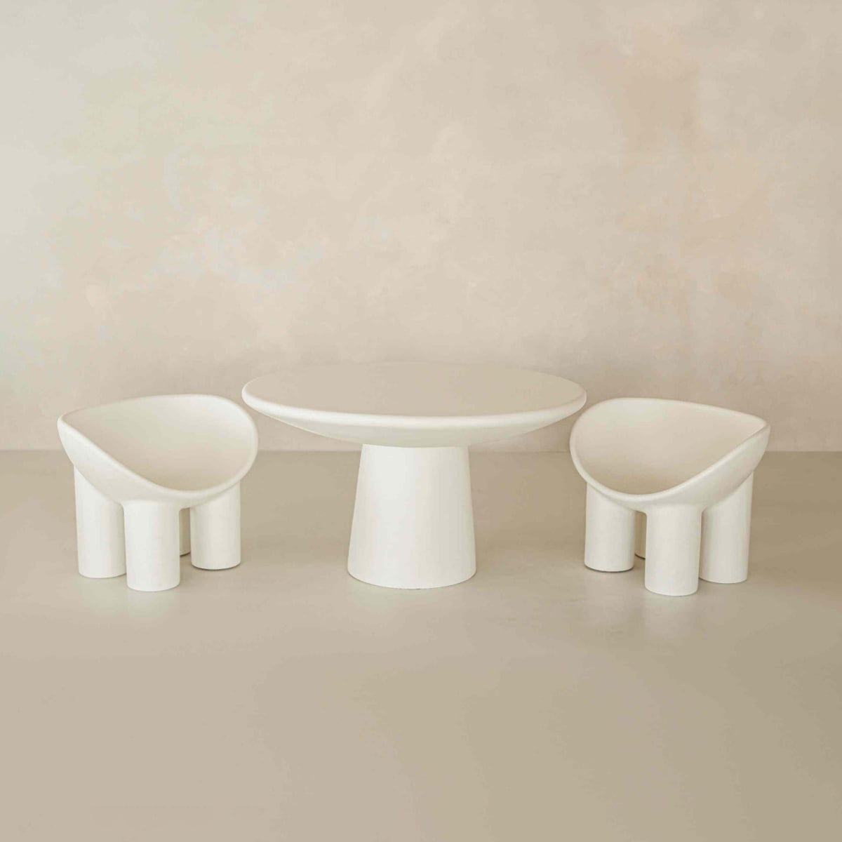 Minimalist set of white furniture with a round table and two matching Arrio Chairs, showcasing thick cylindrical legs and smooth curved seats, arranged against a neutral beige background for a modern sophisticated look.