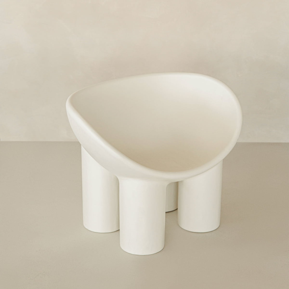 The Arrio Chair features a modern white design with a curved bowl-shaped seat and four thick cylindrical legs, creating an avant-garde statement against a neutral backdrop.