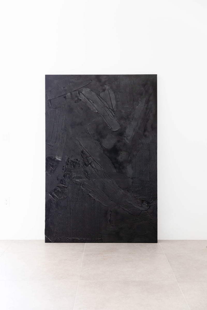 Art By Suzette Newman: A large, rectangular black textured canvas with abstract shapes and raised patterns leans against a white wall on a light beige tiled floor, highlighting unique handcrafted creations in varying black tones.